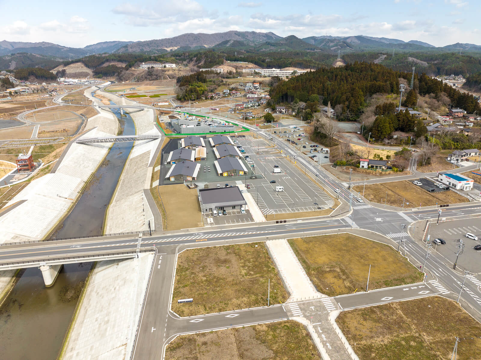 川の右岸に沿って整備された道路や駐車場のある建物群があり、手前には橋が川を横断しており、周囲には舗装された広い道が碁盤目状に伸びている復興地域の昼間の風景を高台から定点カメラで撮影した写真