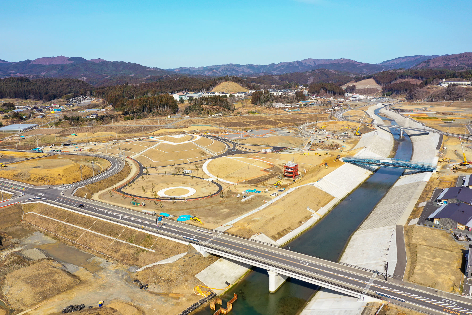 川を横断する橋を中心に、複数の道路や円形の構造物が広がり、建設車両や機材が点在する開発中の建設現場を空から見下ろした、周囲に丘や山が広がる地域の写真