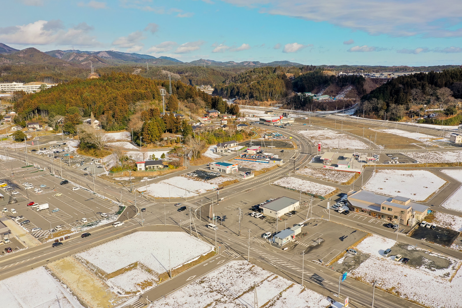 上空から撮影したもので、周囲には丘や森林が広がり、雪が積もった畑や道路が点在し、複数の交差点や駐車場が見え、電柱と電線が町中に張り巡らされており、空は雲が少し浮かぶ晴れた寒い日の様子を写した写真
