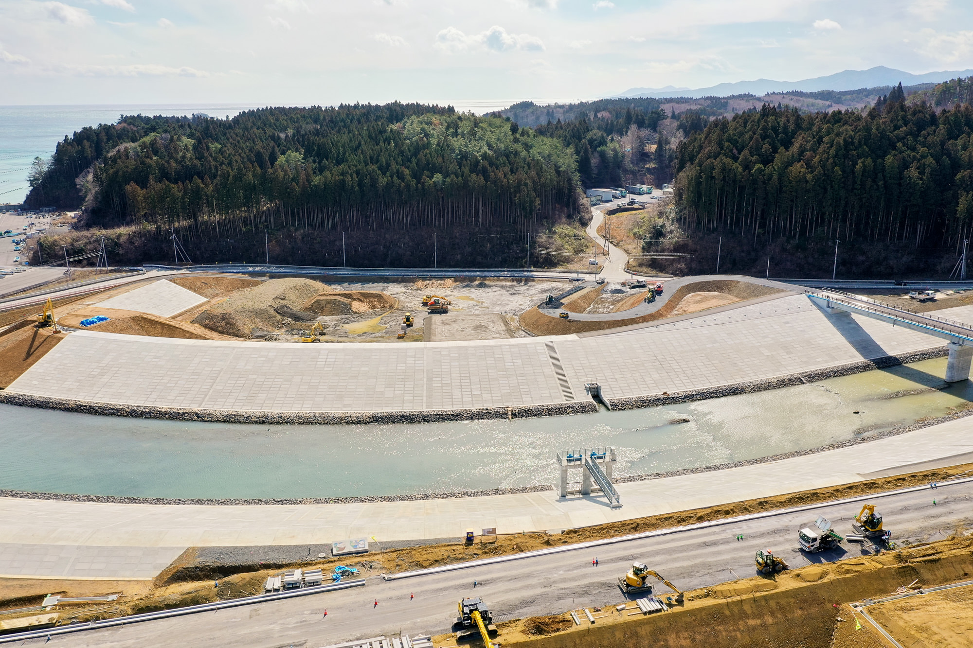 川が中央を流れる広大な工事現場で、両岸にはコンクリート構造物が設置されており、治水施設の一部と見られ、周囲には森林が広がり、建設車両や機材が点在し、背景には丘や山が見える治水工事現場の写真