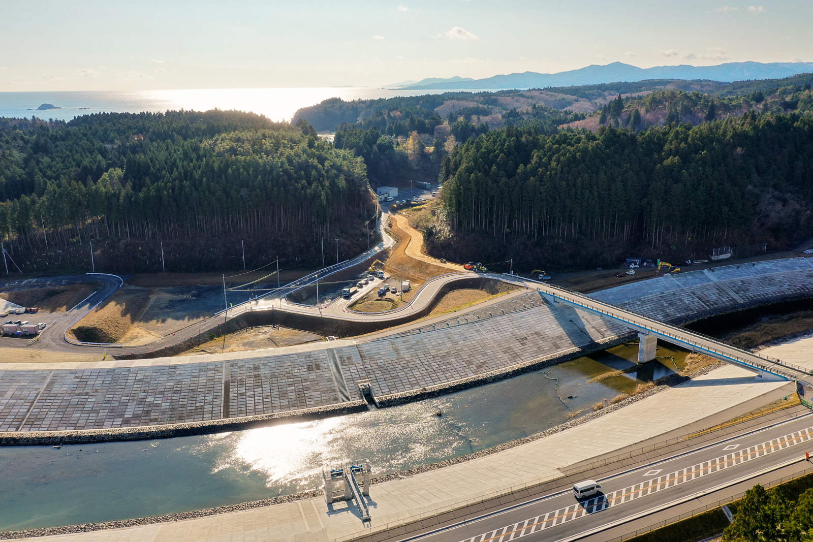川と橋を中心とした風景を上空から撮影した画像で、川はコンクリートの護岸に沿って流れ、並行して道路が走り、車両が見られ、橋が川を横断して両岸をつなぎ、背景には森林に覆われた丘が広がり、地平線の向こうには水面が輝き、太陽が昇るか沈む様子が写された川と橋のある風景の写真