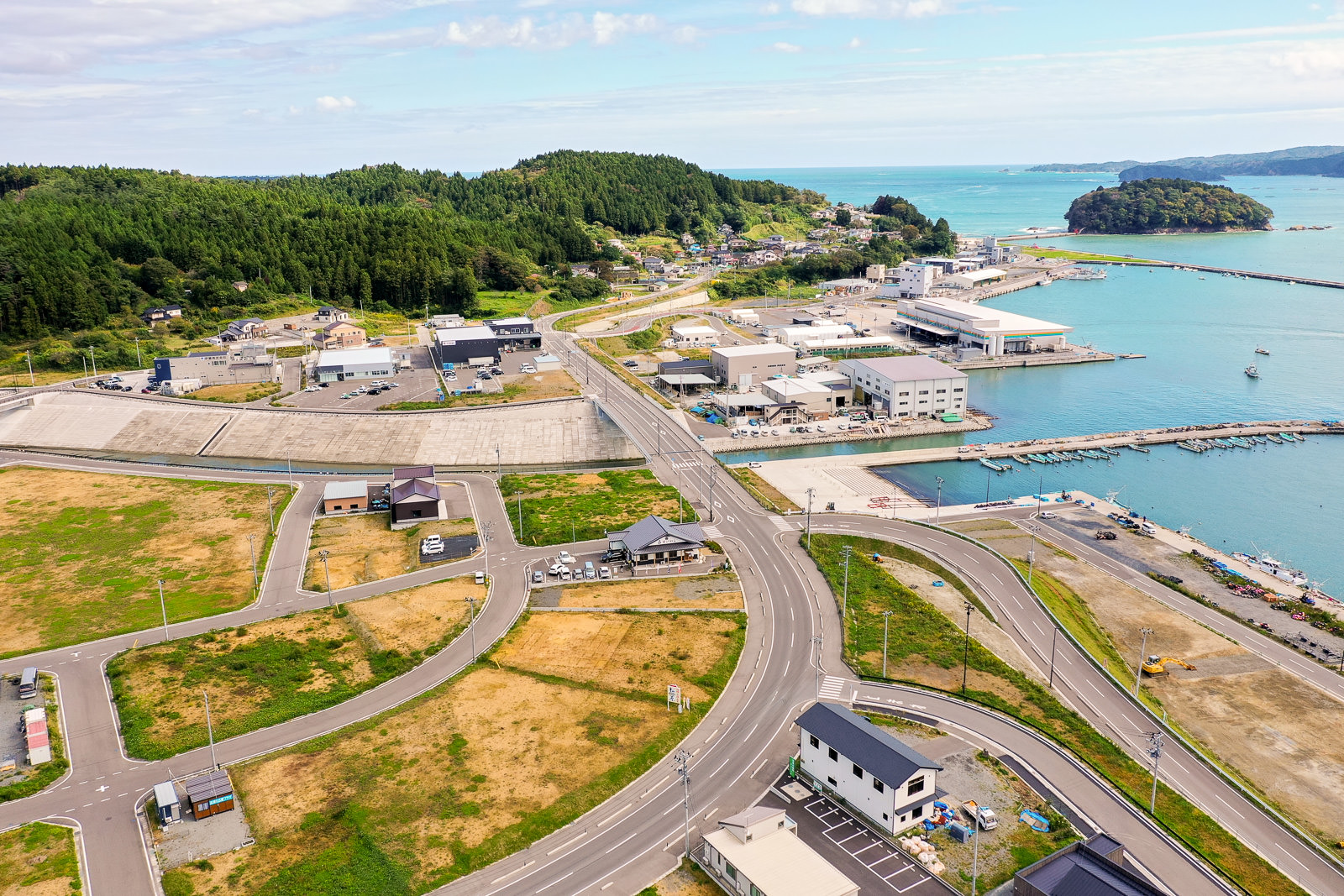 住宅と工業施設が混在する沿岸の町を空から捉えた風景で、複数の道路が交差しながら家屋や倉庫へと続き、海辺には桟橋や船が見え、周囲には緑の丘や森林が広がる港町の写真