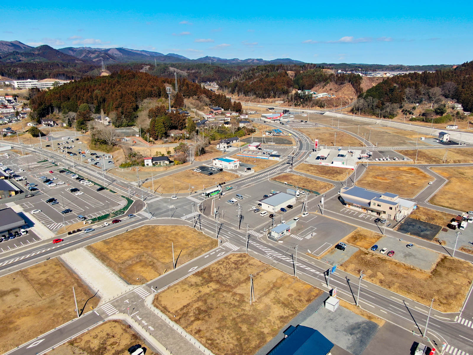 上空から見下ろした風景が広がっており、複数の道路や交差点、建物が点在し、草地や舗装された空き地も多く見られ、背景には森林地帯と遠くに広がる丘や山々が写っている写真