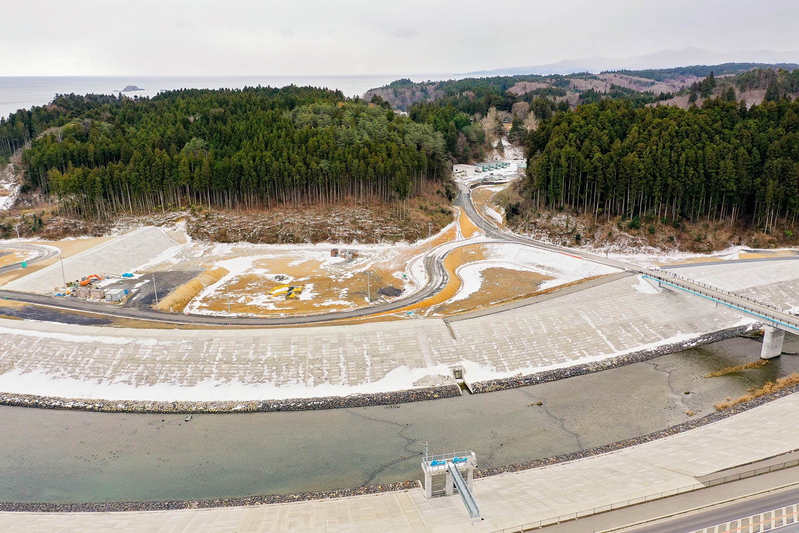 河川の両岸を高くコンクリートで固められた護岸が伸びており、川の上には橋がかかり、その奥には山々と積雪した道路が続く人里離れた山間のインフラ整備地域の昼間の風景を高台から定点カメラで撮影した写真