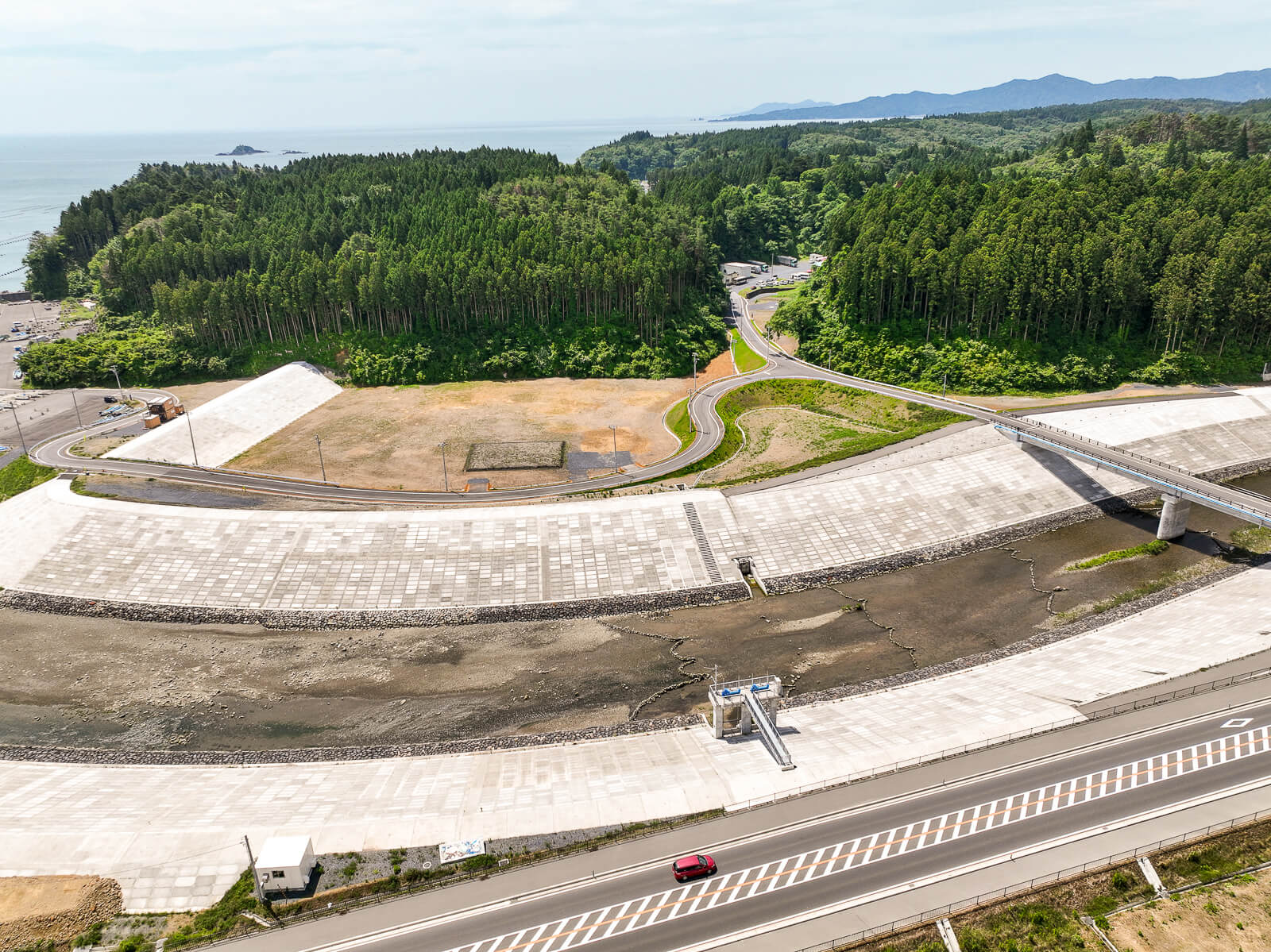 防波堤の内側に整備され舗装された道路と空き地があり、その背後には緑豊かな丘陵地帯が広がっている昼間の風景を高台から定点カメラで撮影した写真