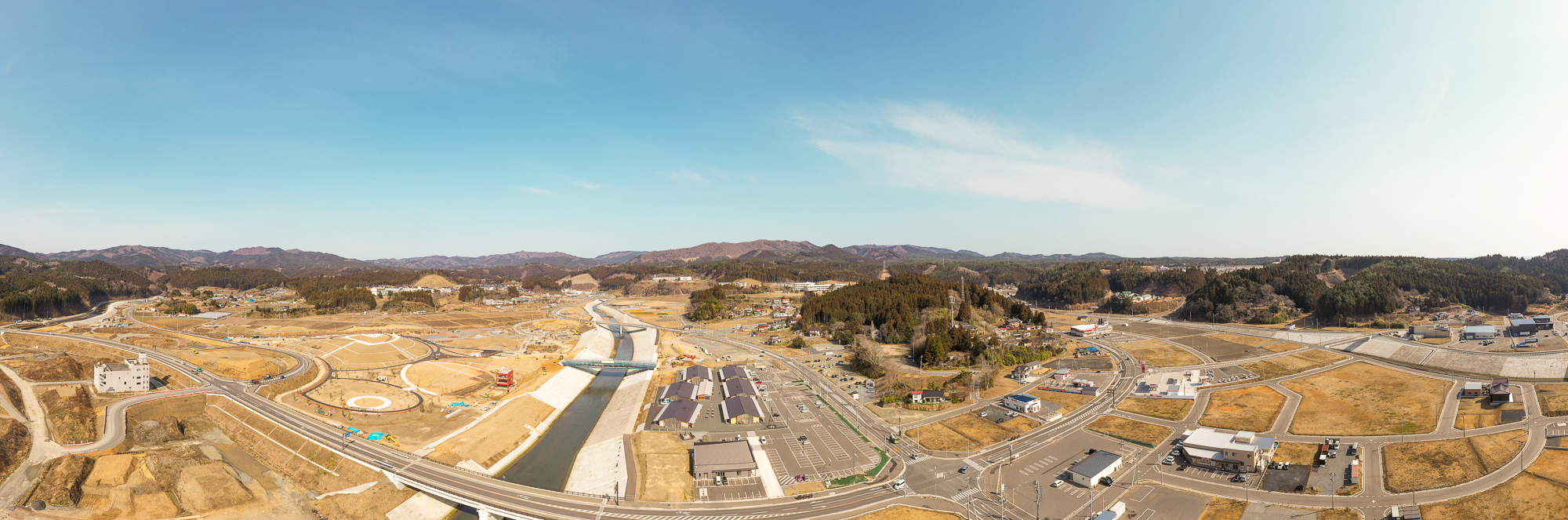 広い空の下、山々に囲まれた地域の中央を川が流れ、その周囲に道路や建物が点在している様子を広く見渡せるパノラマ風景の写真