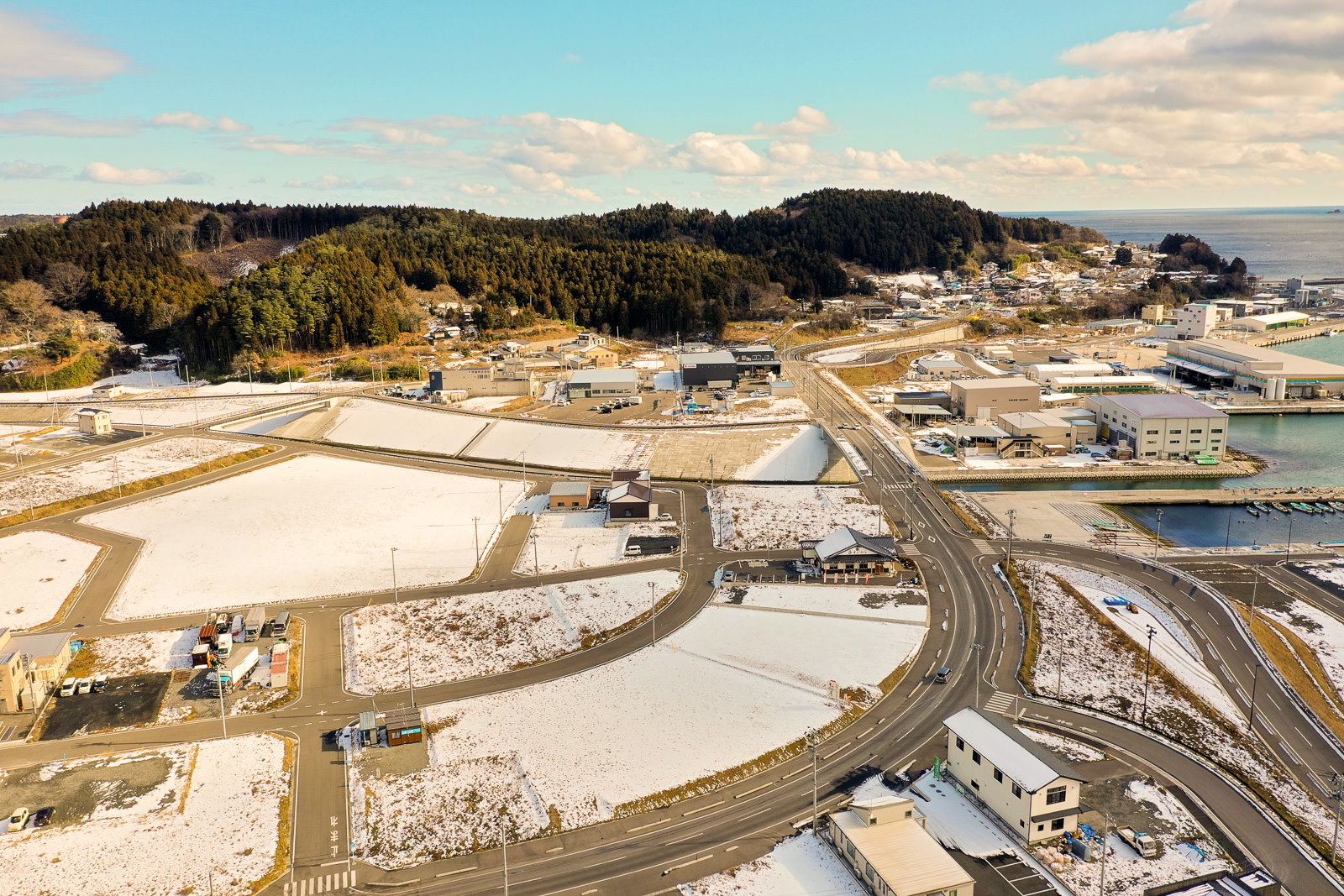 雪が積もった畑や道路、建物が広がる海沿いの町を上空から撮影したもので、住宅と工業施設が混在し、背景には木々に覆われた丘があり、右側には海が広がっており、空は雲が少し浮かぶ晴れた冬の日の様子を写した海辺の町の写真