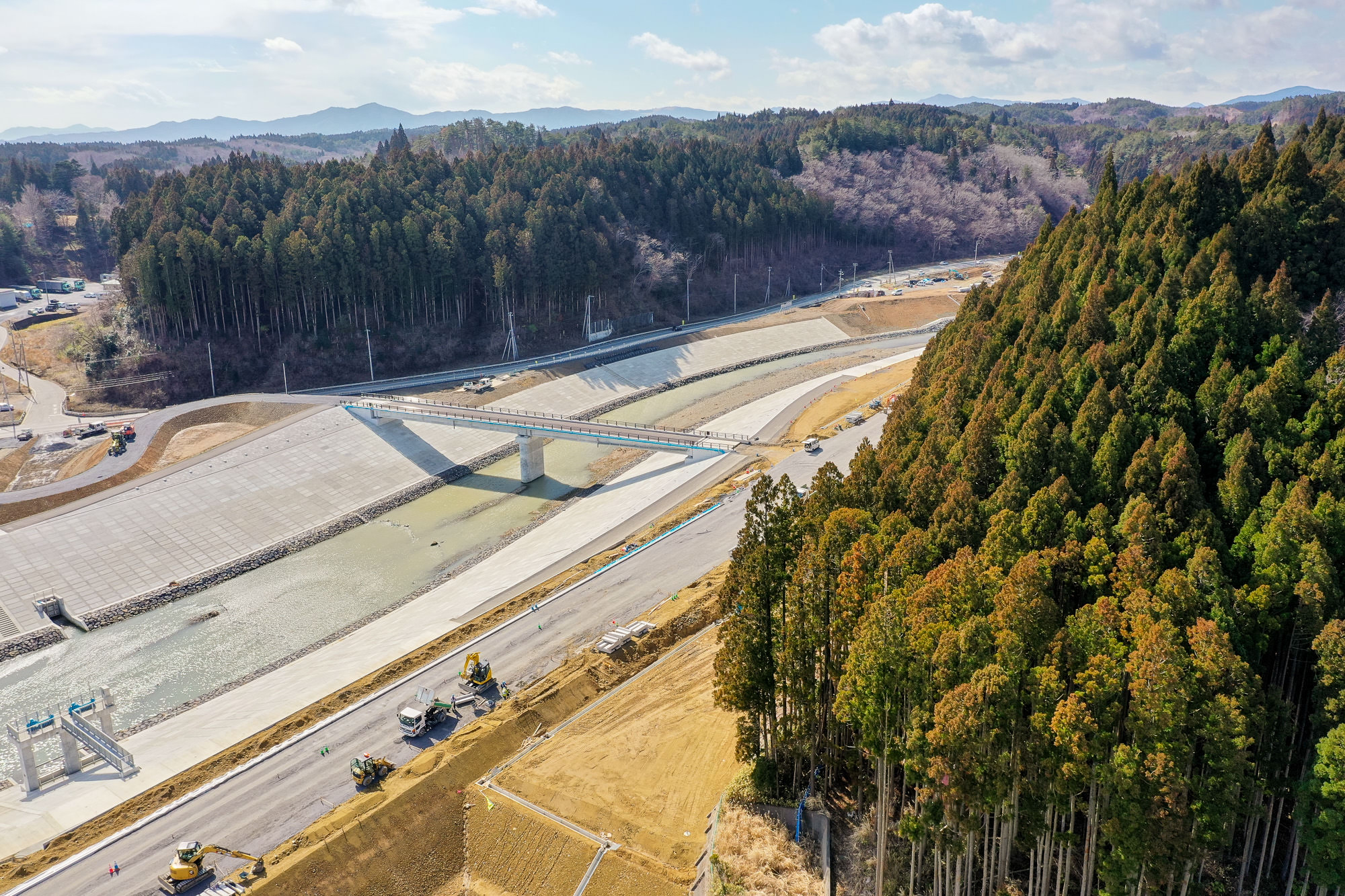 中央に水が流れる幅広いコンクリート製の水路が通り、その上に橋が架かり、両側には道路と建設車両が見られ、周囲は背の高い木々が密集する森林に囲まれ、背景には山々が広がる、部分的に雲が浮かぶ空の下の森林地帯の工事現場の写真