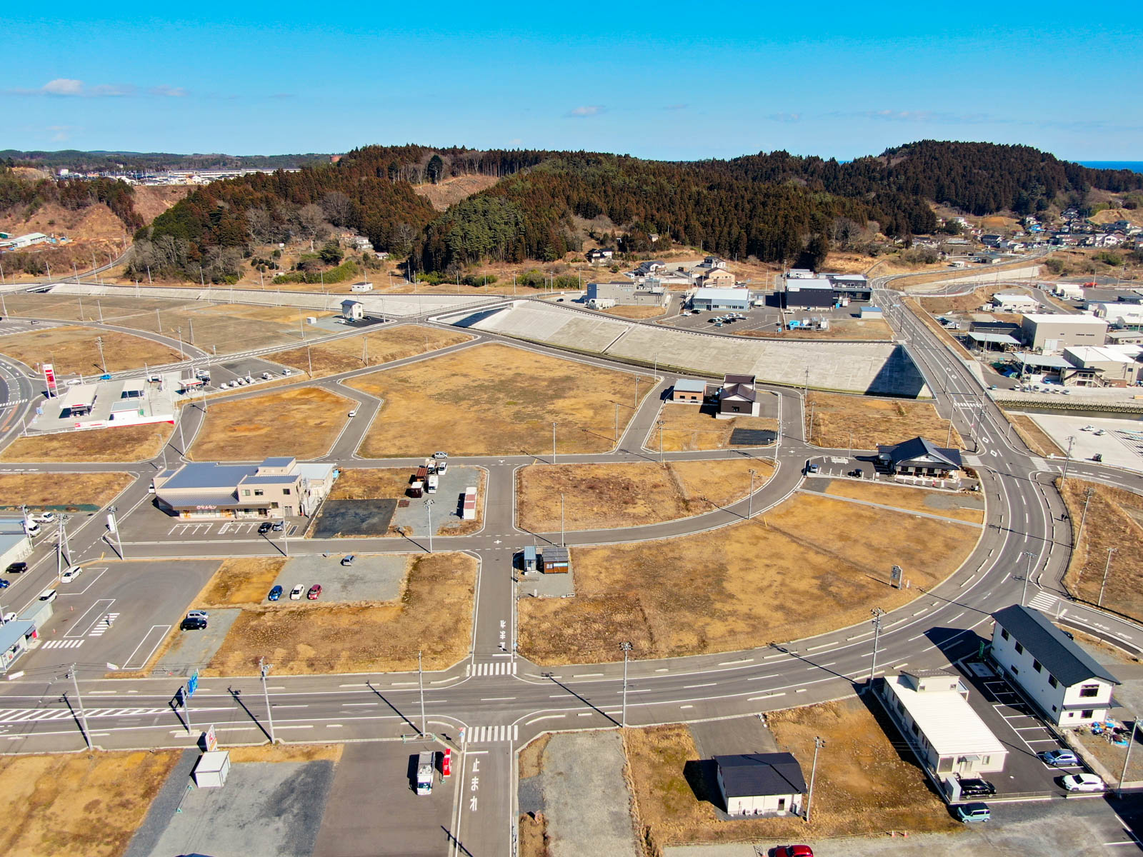 郊外の地域を上空から見下ろした風景が広がっており、道路網と区画された土地が整然と配置され、建物や車両はまばらに点在し、交差点や横断歩道、駐車中の車も見られ、背景には木々に覆われた丘が広がり、空は澄んだ青空が広がっている写真