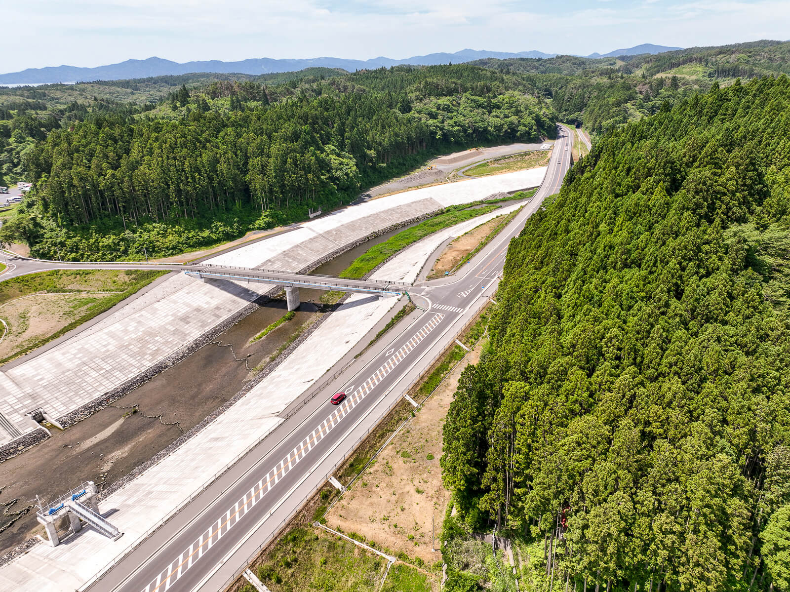 道路と交差する川の防波堤の脇に架かる橋が道路を跨ぎ、その横に緑濃い森林が広がっている昼間の風景を高台から定点カメラで撮影した写真