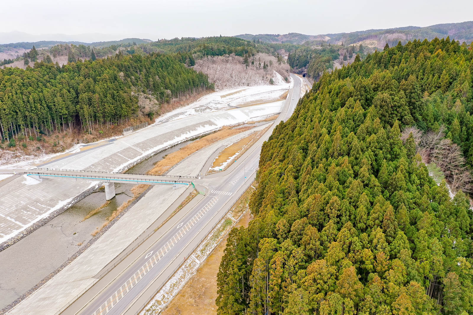 橋が架かる川沿いの道が右奥へ向かって延びており、道の両側は雪が積もった林に囲まれ、整備されたインフラと自然が共存する山あいの昼間の風景を高台から定点カメラで撮影した写真