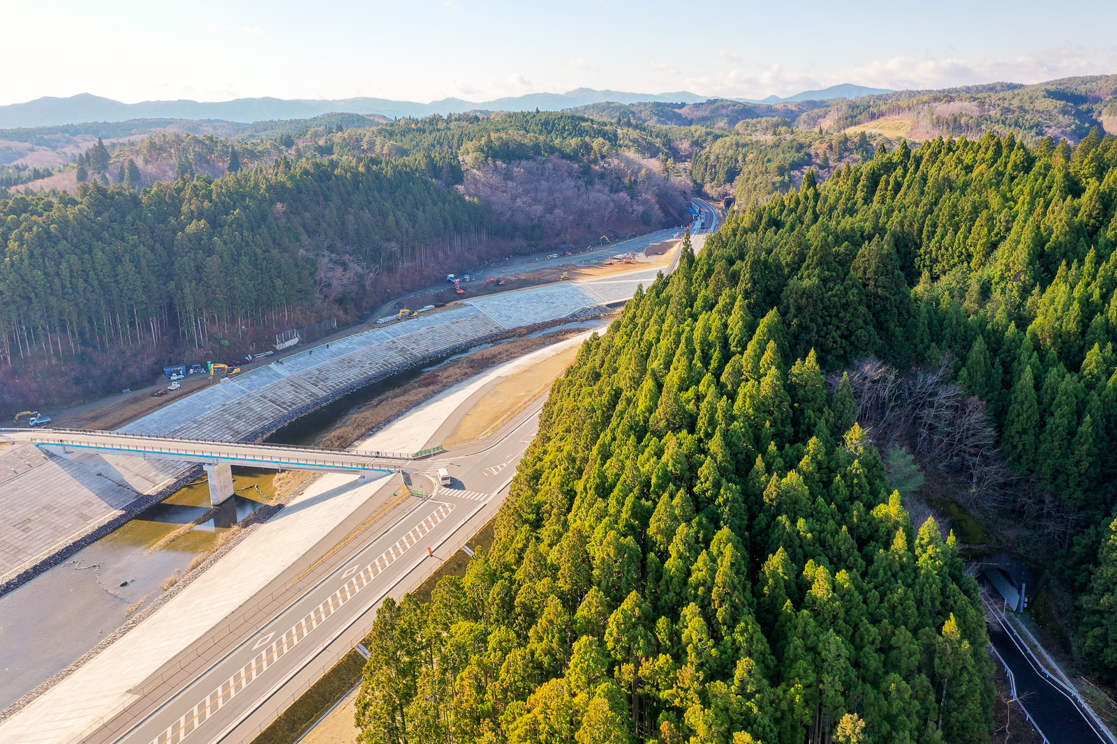 右側に緑豊かな森林が広がり、左側には造成中またはインフラ整備中と思われる広い開けた土地があり、その上を橋が横断して両側をつなぎ、背景には木々に覆われた丘が連なり、遠くには山々が見え、空は晴れている自然と開発が共存する風景の写真