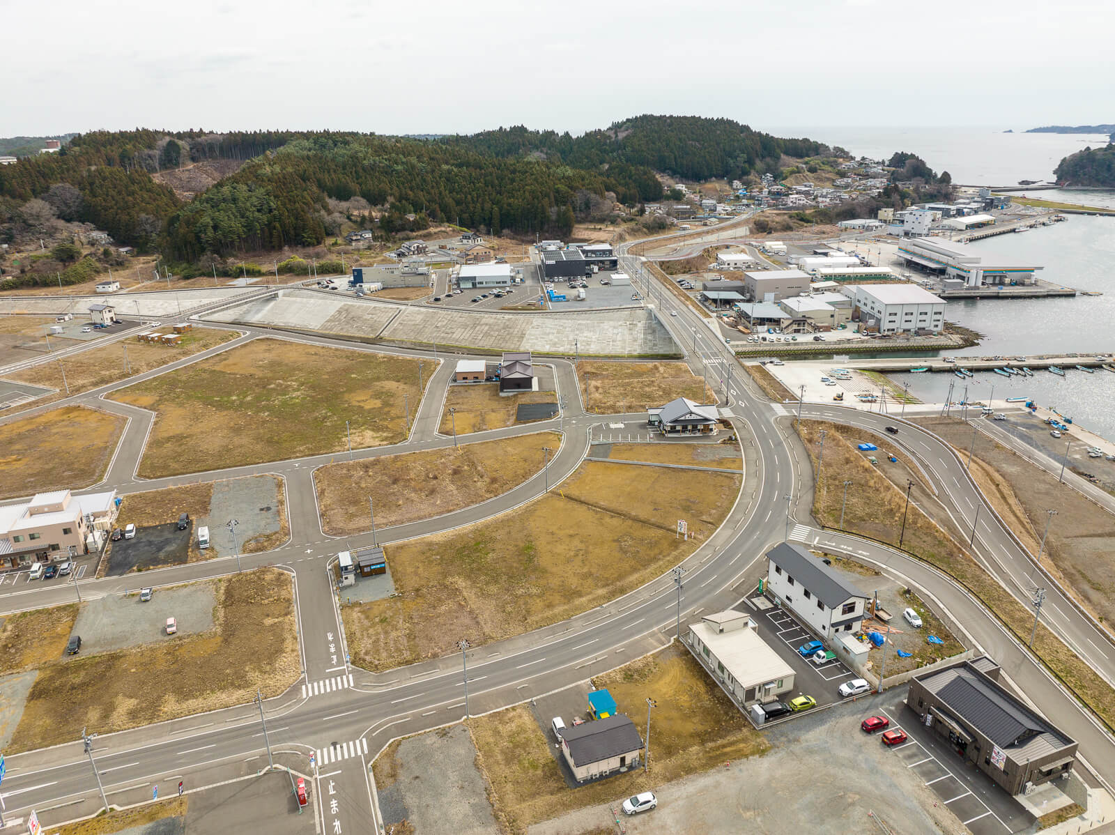 カーブを描く道路の周囲に数軒の建物と整備された区画が並び、背景には港の建物群や海が見え、漁港へ続く道と再開発エリアの境目が見える、港近くの市街地と海への接続部の昼間の風景を高台から定点カメラで撮影した写真