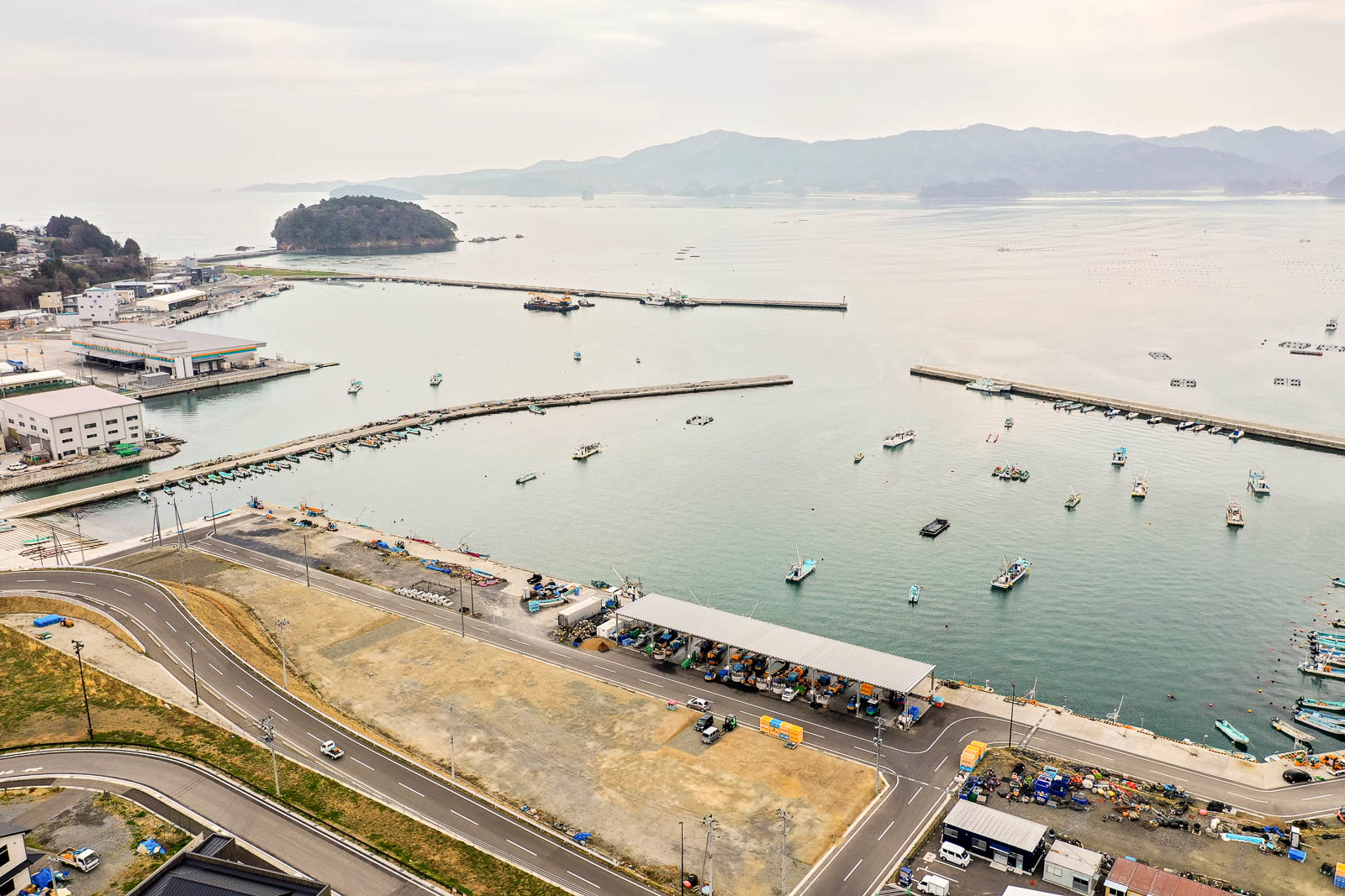 海に突き出た防波堤に囲まれた港に複数の船が停泊し、海岸沿いには建物や道路、駐車場などのインフラが整備され、背景には山々と海の向こうに島が見える様子を上空から捉えた風景の写真