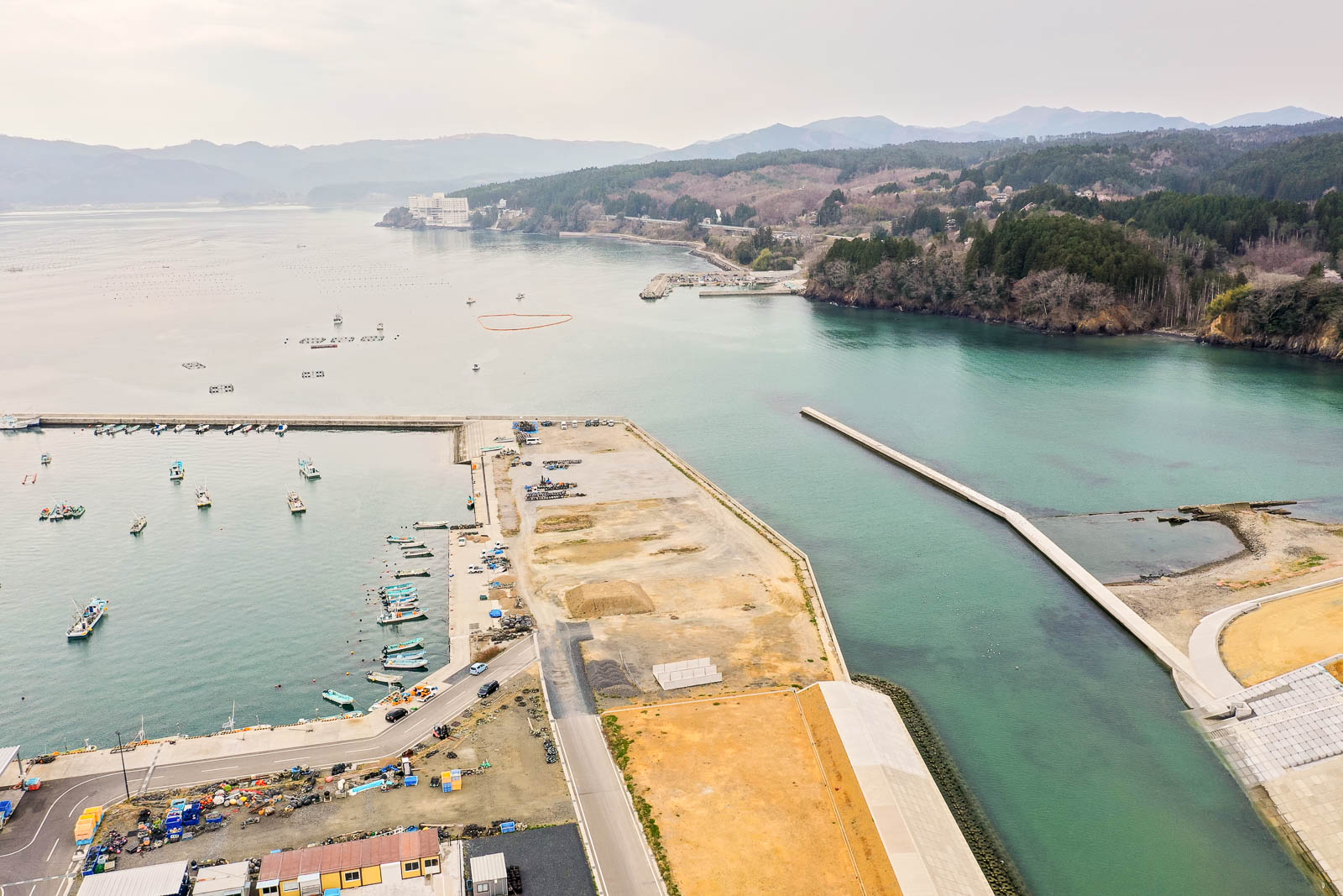 港に複数の船が停泊し、周囲には建物と広い空き地が広がり、穏やかな海と背景に見える丘や山々が調和した様子を上空から捉えた風景の写真