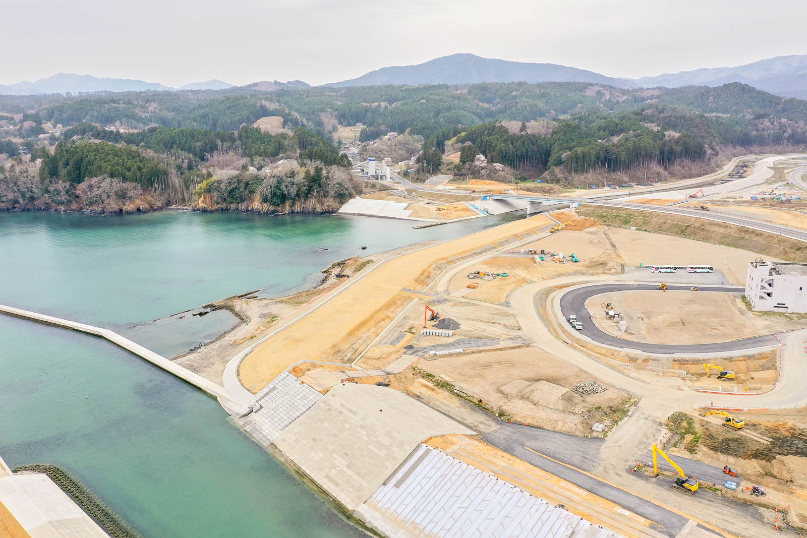 左側に水辺とコンクリートの護岸が広がり、複数の道路や整地された土地、重機が点在する海沿いの建設現場を上空から捉え、背景には森林に覆われた丘や山々が広がる風景の写真