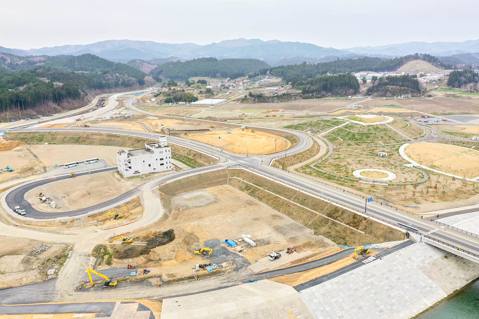 複数の道路といくつかの建物、さまざまな建設車両が点在する広大な建設現場を上空から捉え、周囲には丘と森林が広がる風景の写真
