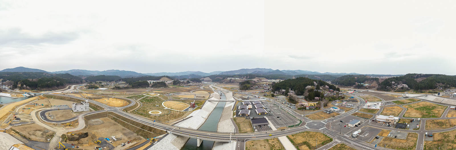 広大な開発地を上空から捉え、複数の道路や橋、建設中の建物が点在し、将来的な造成が予定されている区画も見られ、周囲には緑と木々が散在し、背景には山々が広がる風景の写真