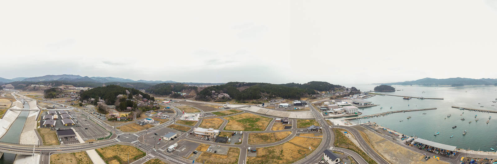 港に複数の船が停泊し、海沿いに建物が並ぶ町を上空から捉え、道路が町を縫うように走り、背景には山々が広がり、開発された地域と緑の空間が共存する様子が見える風景の写真
