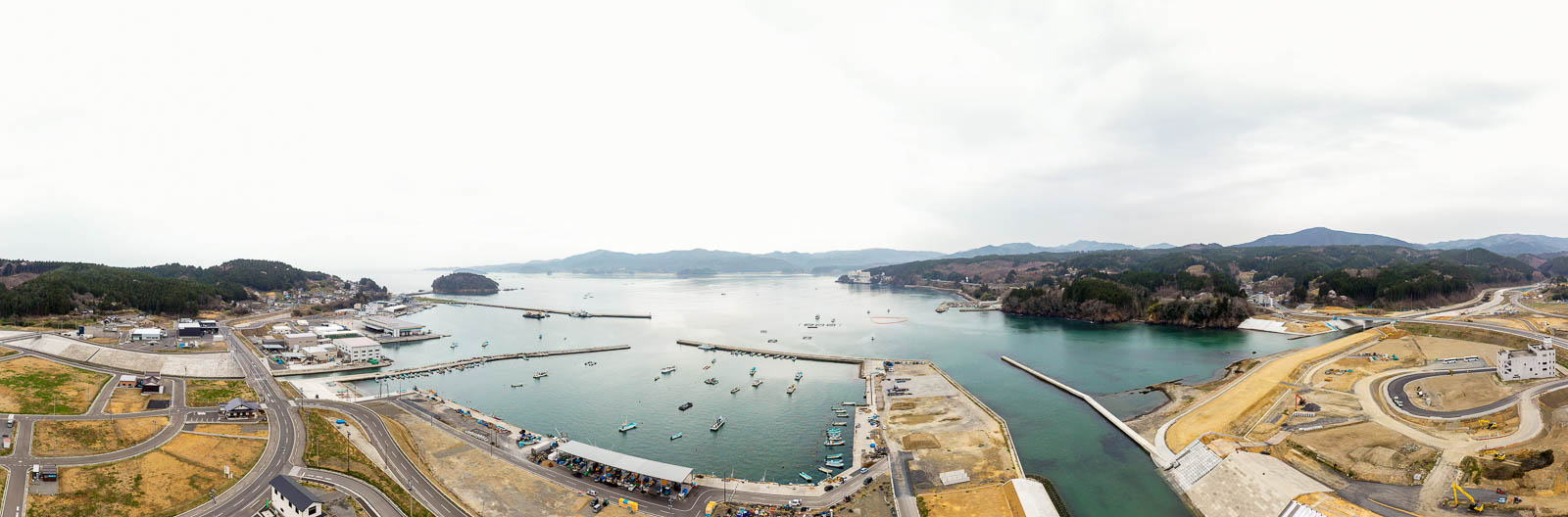 海沿いの地域を上空から捉え、港には複数の船が停泊し、岸辺には建物やインフラが並び、道路や駐車場、緑地が点在し、背景には木々に覆われた山々が広がり、空は曇っている風景の写真