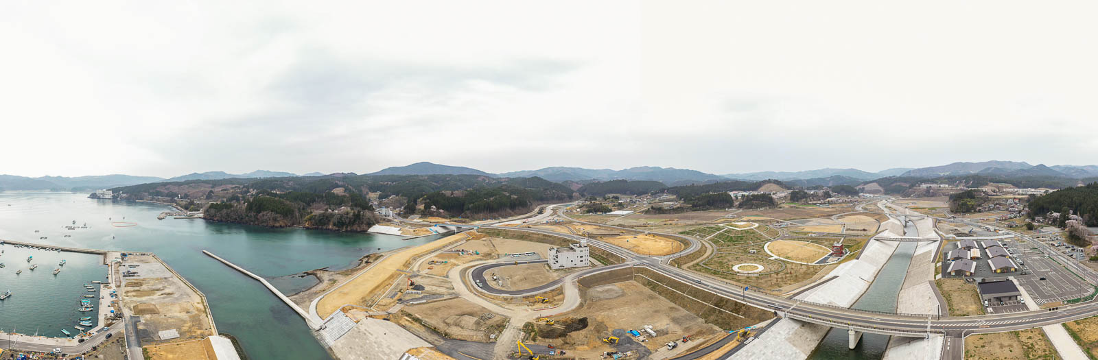 開発または再整備中の海岸地域を上空から捉え、複数の工事現場や道路、インフラが広がり、港には船が停泊し、背景には丘や山々が連なる風景の写真