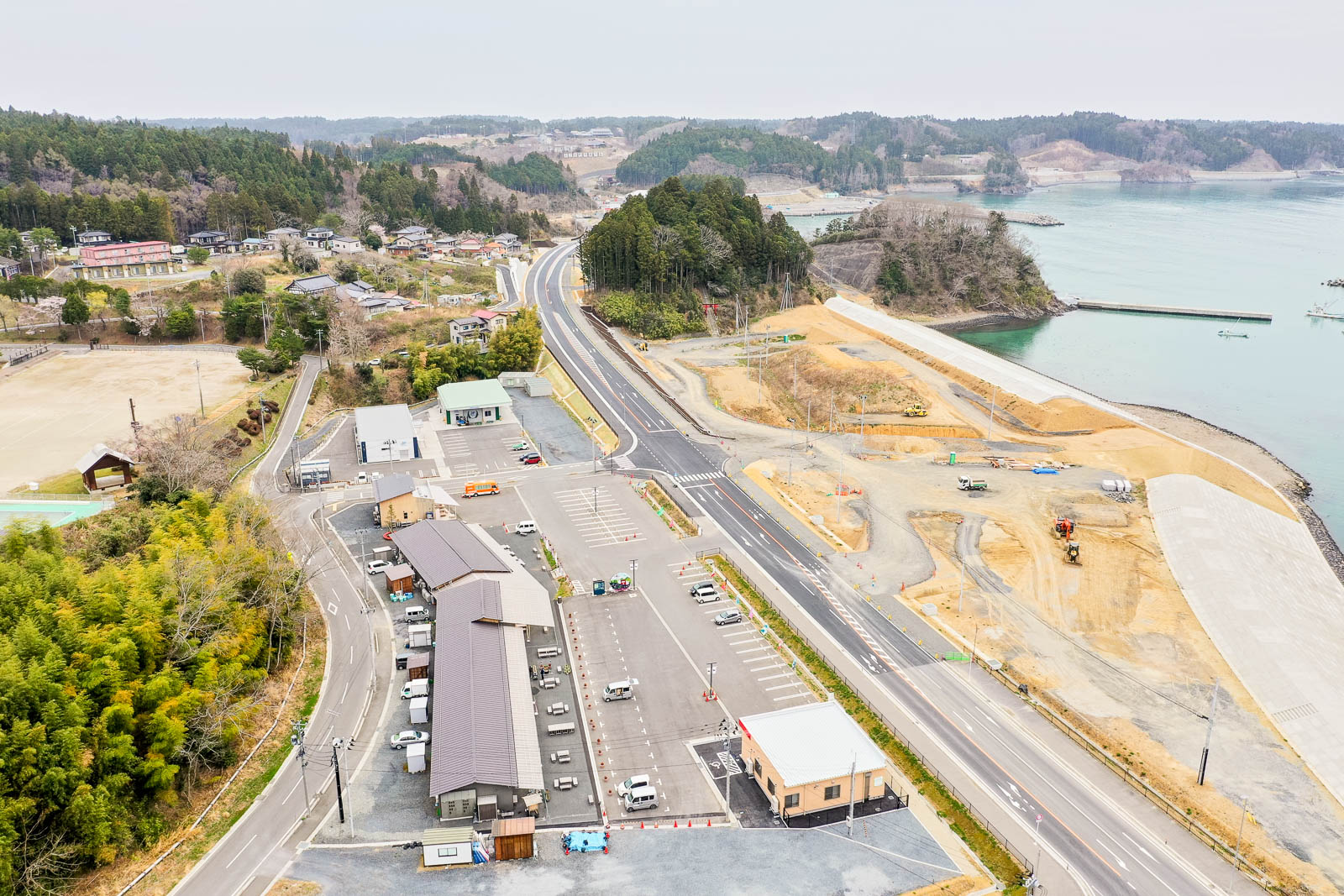 海岸線に沿って道路が走り、その両側には建物や駐車場が並び、車が停まっている様子が見え、自然の風景と開発地が混在し、右側には海が広がり、海辺では工事や造成が行われている様子を上空から捉えた風景の写真