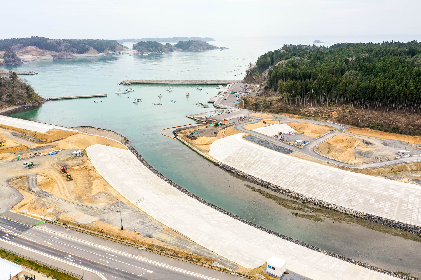 港に複数の船が停泊し、海岸沿いにはコンクリート構造物や道路が整備され、建設機械も見られ、背景には森林と遠くに小さな島々が広がる様子を上空から捉えた風景の写真