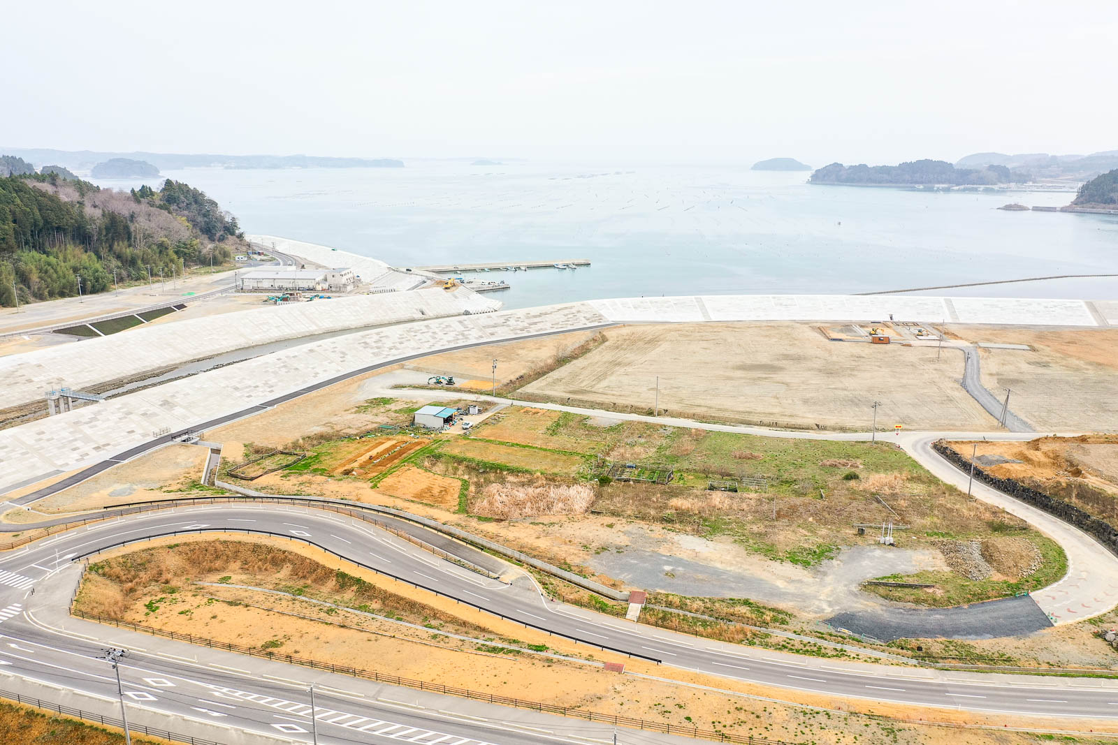 広い空き地や開発地と未開発地が入り混じった海岸地域を上空から捉え、曲がりくねった道路が走り、遠くには水平線まで広がる海が見える風景の写真
