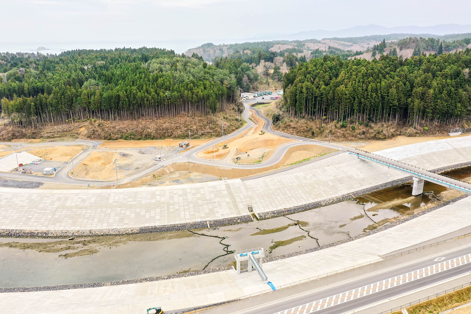 中央に大きなコンクリート構造物が走り、少量の水が流れる水路のような形状をしており、その両側には道路や歩道が広がり、周囲には密集した森林と背景に丘が広がる様子を上空から捉えた風景の写真