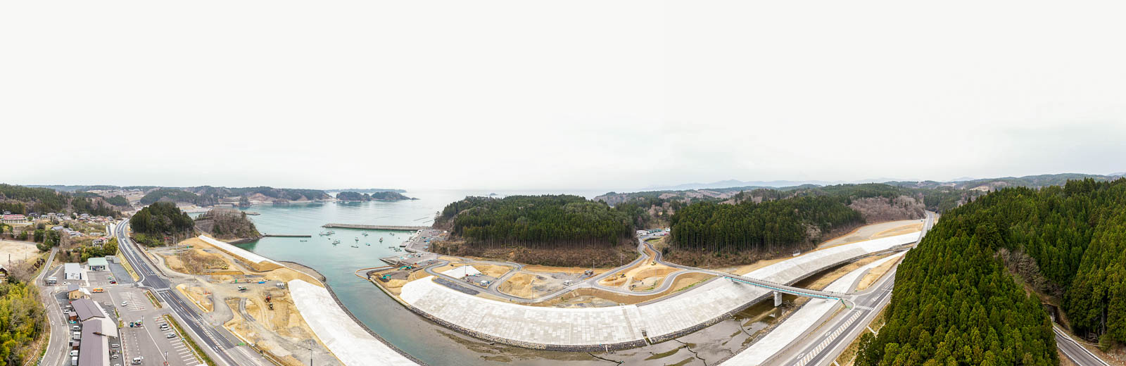湾に面した海岸地域を上空から捉え、森林に覆われた丘に囲まれ、海岸沿いには防波堤や堤防が整備され、湾内には船が停泊し、左側には建物や構造物が点在する風景の写真