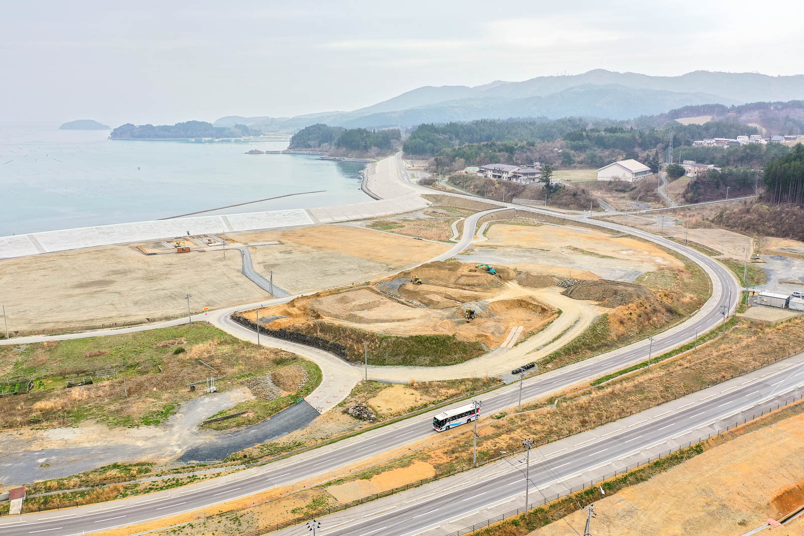 曲がりくねった道路と海沿いの建物、開発中の土地が広がり、背景には山々がそびえ、陸と海の両方を見渡せる高所から撮影された風景の写真