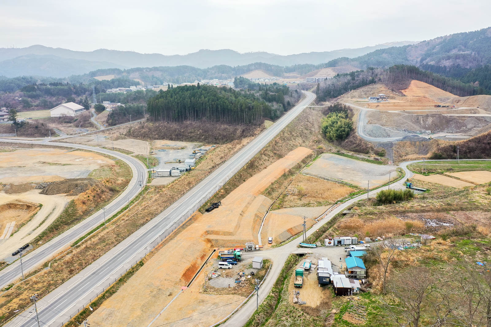 丘と森林に囲まれた風景の中を道路が走り、その両側には工事現場が広がり、車両や重機が点在し、背景には山々と曇り空が広がる様子を捉えた風景の写真