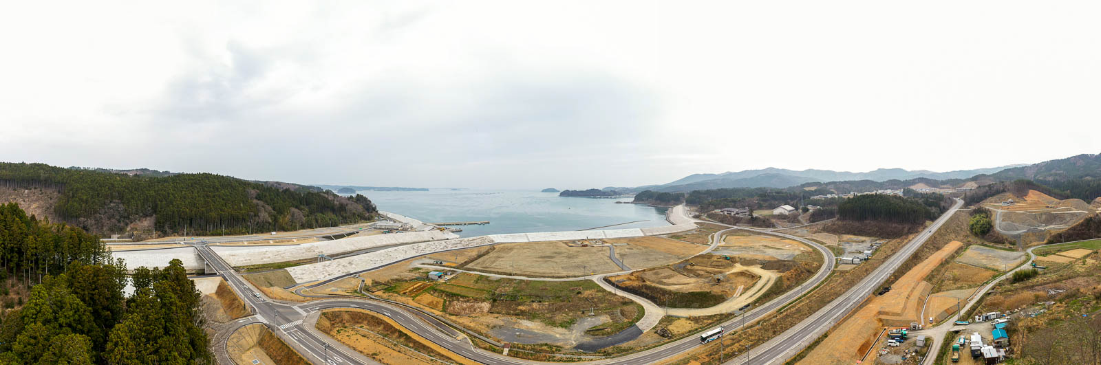 海岸線に沿って広がる町並みと自然が入り混じり、曲がりくねった道路や開発中の土地、緑の斜面が点在し、遠くには海と山々が見える広大な景色の写真