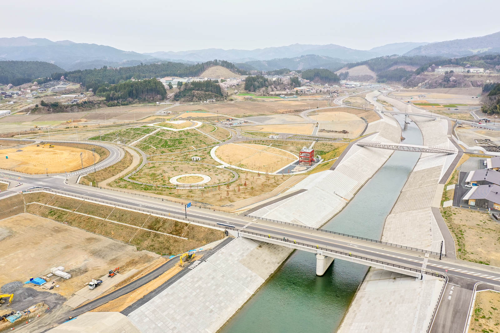 コンクリートで整備された川が複数の橋が架かり、その周囲には開発中または整備された土地が広がり、道路や歩道、緑地が点在し、背景には丘と森林が広がる様子を上空から捉えた風景の写真