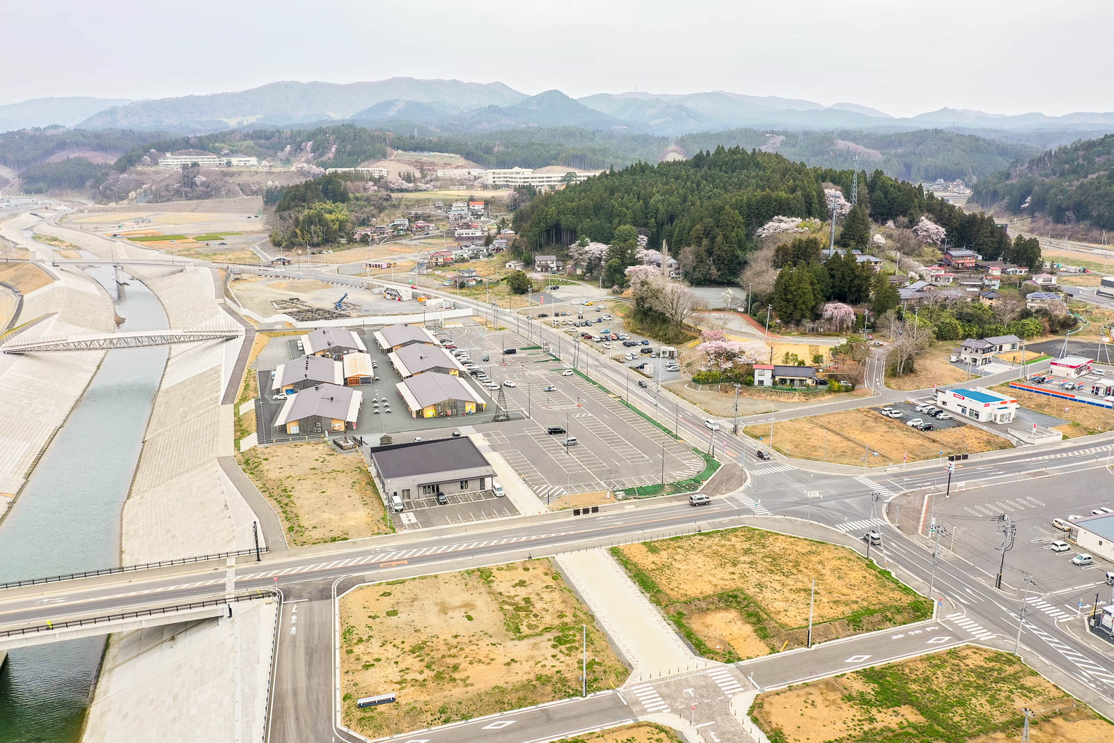 複数の建物と道路が広がる小さな町または村を上空から捉え、川が町の中を流れ、交差点や駐車場が点在し、周囲には丘と森林、背景には山々が広がり、桜の木が咲いている様子も見える風景の写真