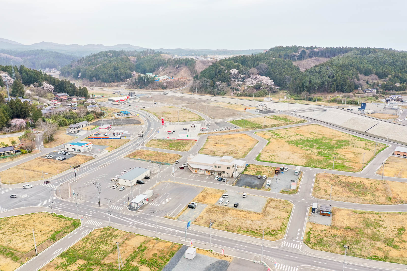 碁盤の目のように整備された道路に沿って建物や駐車場、車両が点在し、草地や木々が混在する半都市型の地域を上空から捉え、背景には丘と森林が広がる風景の写真