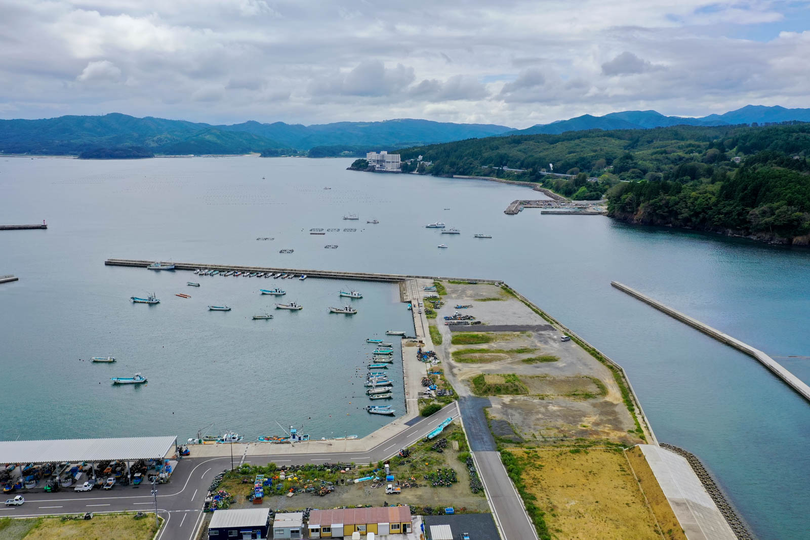 防波堤に囲まれた港に小型漁船が並び、岸壁には漁具や作業車両が整然と配置され、奥には山々が広がっている漁港の写真