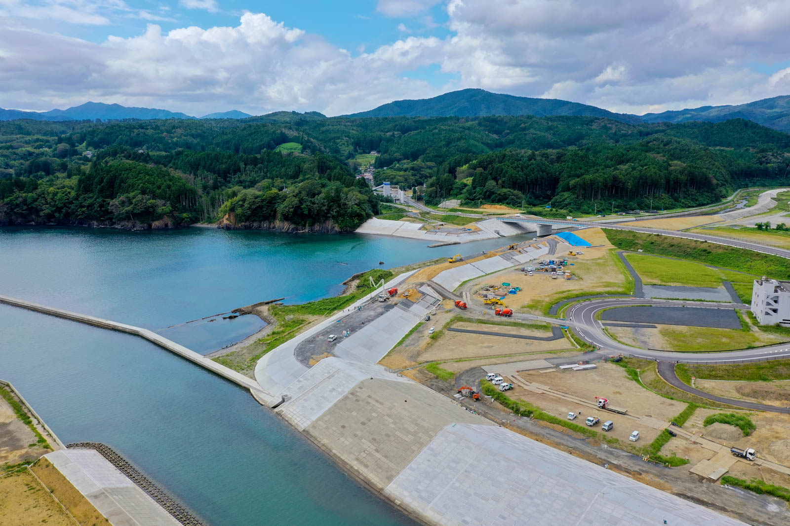 海と森に囲まれた小さな湾に面して大規模な防潮堤と工事中の道路が整備され、クレーンや工事車両が点在する、津波被害からの復興工事中の港湾部の写真