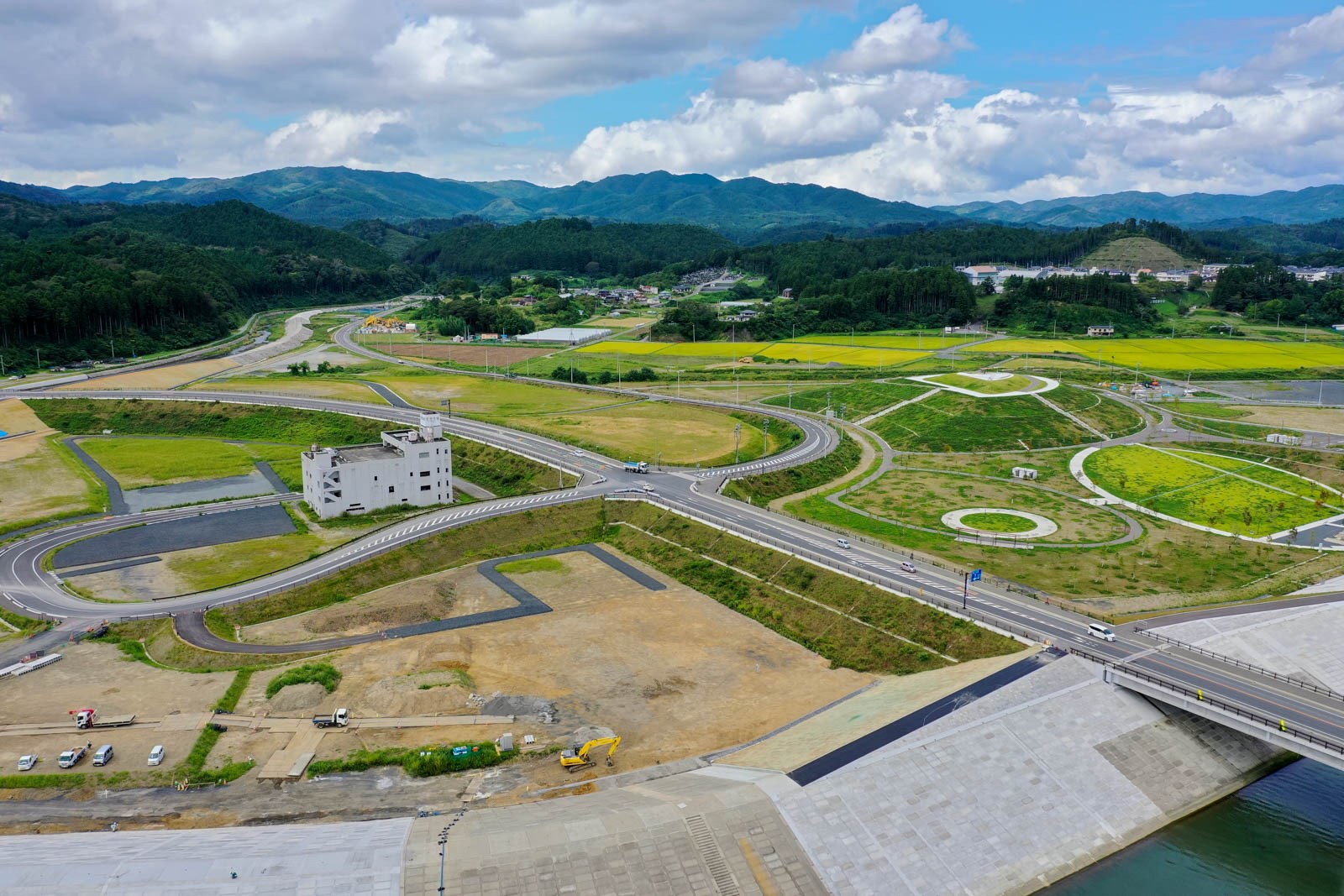 高台から見下ろす形で、複数の交差点と円形の緑地が整備され、中央には震災遺構と見られる白い鉄筋コンクリートの建物が残り、その周囲に新しい道路が伸びている、復興エリアの写真