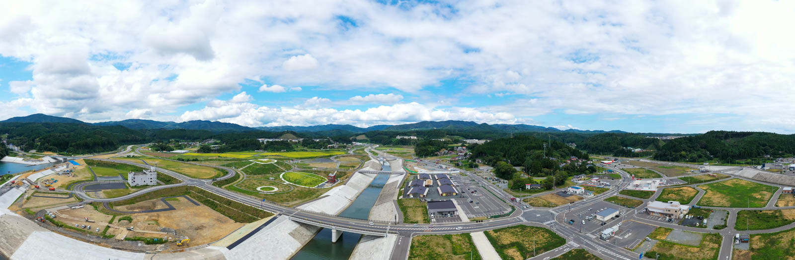 堤防の上空から全体を俯瞰しており、整備された河川、道路、緑地、そしてその奥に山並みが広がっている風景の写真
