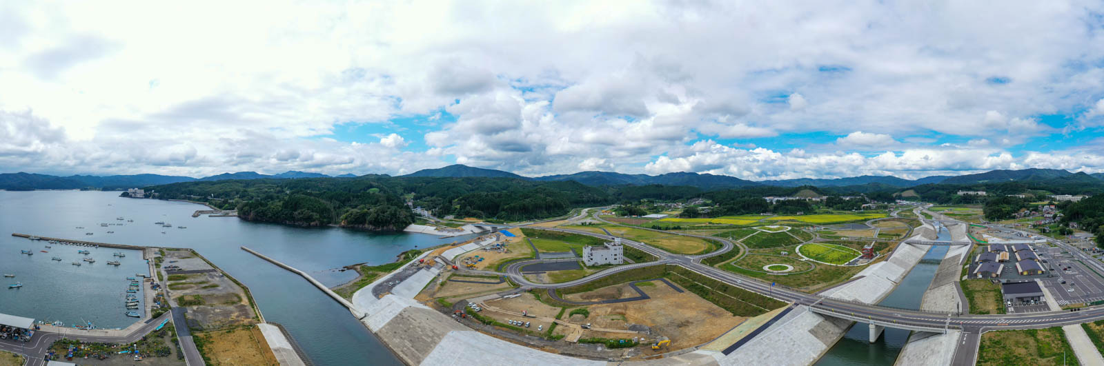 港の防波堤と海岸線に沿って整備された広大な防潮堤と道路が湾奥まで続き、山々と青空を背景に復興された町並みが広がっている、湾周辺全体のパノラマ写真