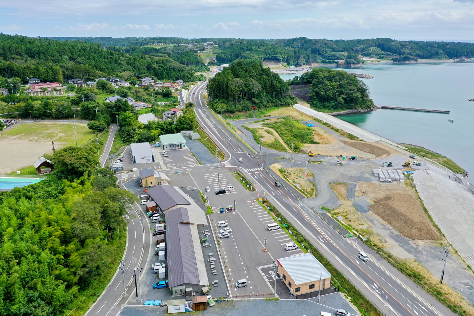 道路に沿って駐車場や建物が並び、その奥に海が広がり、防潮堤や工事中の敷地が確認できる写真