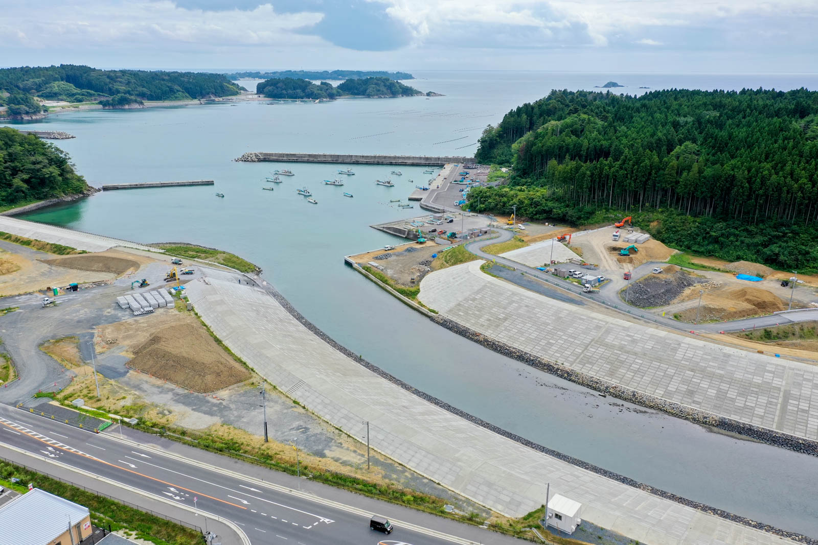 奥に小さな島々が浮かぶ湾内に漁船が並び、防潮堤と整備中の護岸が川を囲むように作られている写真
