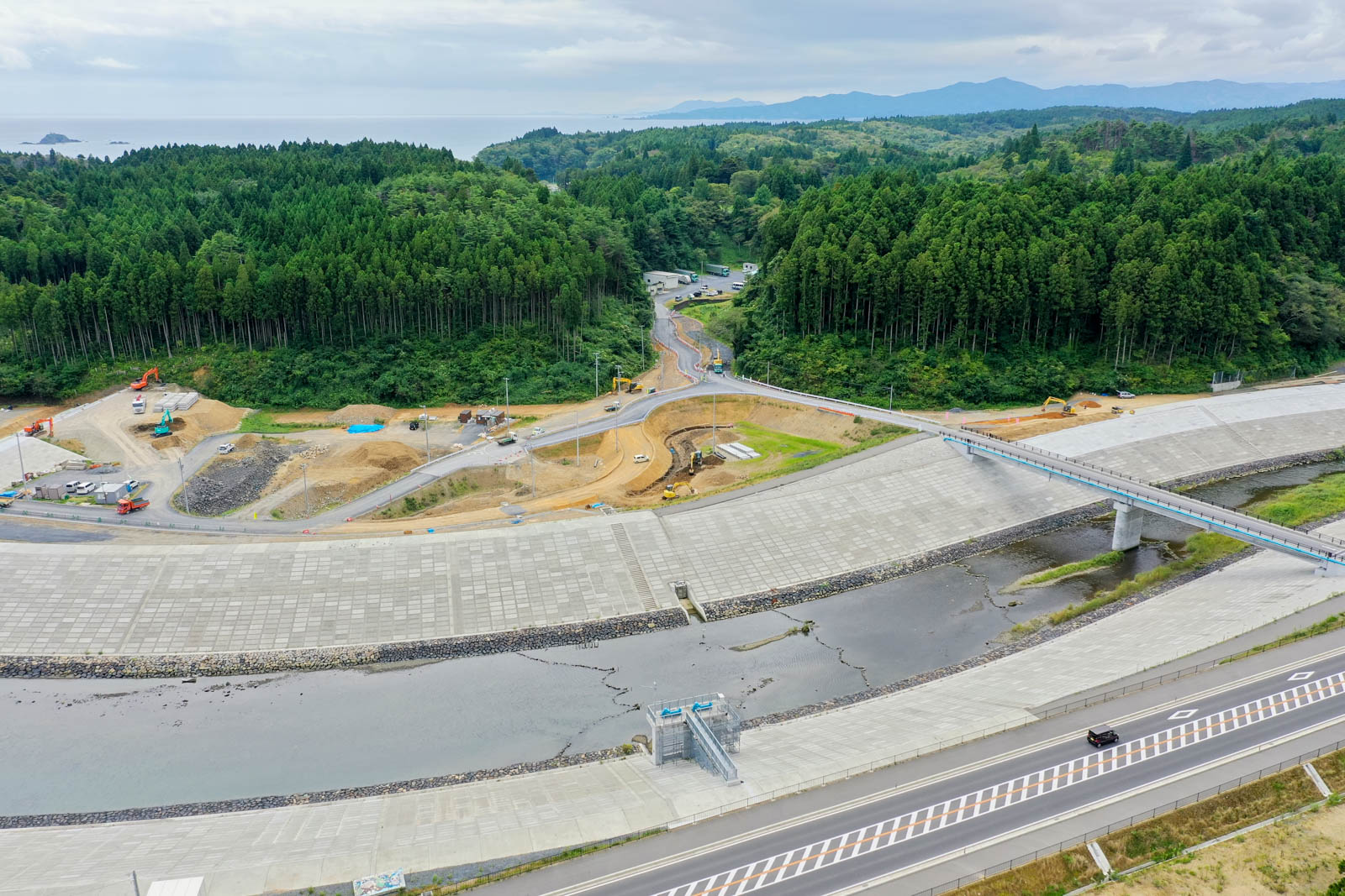 防潮堤の手前に道路と川が見え、奥には林に囲まれた丘陵と建設中の道路が見える写真