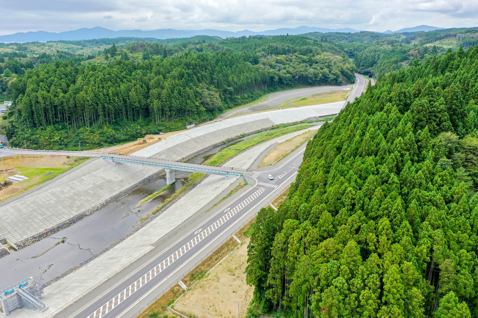 コンクリート護岸の上に架かる歩道橋の奥に、深い緑の森林と曲がりくねった道が山へと続いている写真