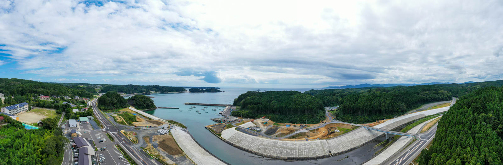 高台から海に向かって撮影されており、中央には防潮堤で仕切られた湾とそこに停泊する漁船が見え、両側を森林に囲まれた港町が広がっている写真
