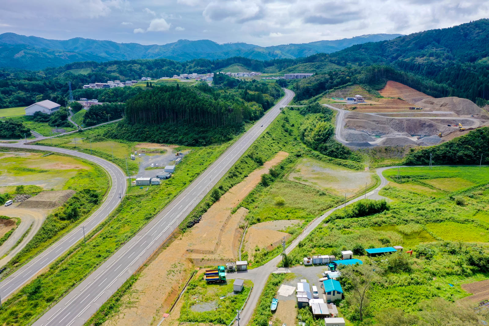 緩やかにカーブを描く道路が山間を走り、その周囲に造成地や工事中の重機、住宅地が点在する地域の写真