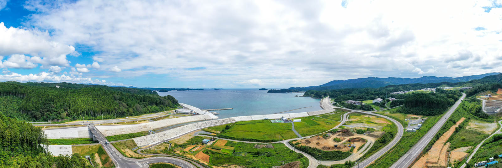 堤防と海岸線が長く続き、奥に湾が広がる海沿いの地域と周囲の緑豊かな山々を広く見渡せる写真