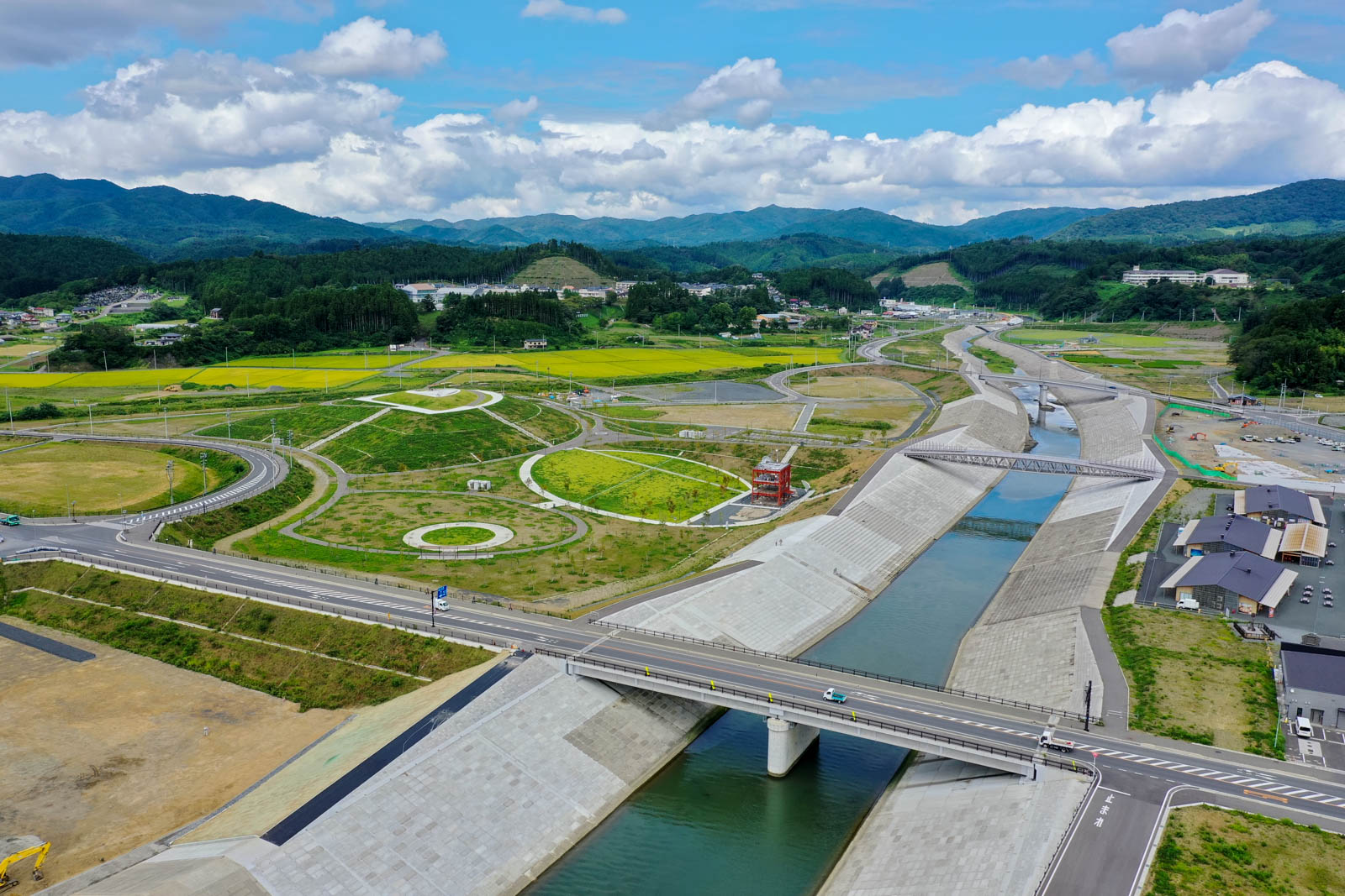 河川と堤防、車道、周囲の公園整備や円形の広場が確認でき、奥に黄色く色づいた田園や山が見える写真