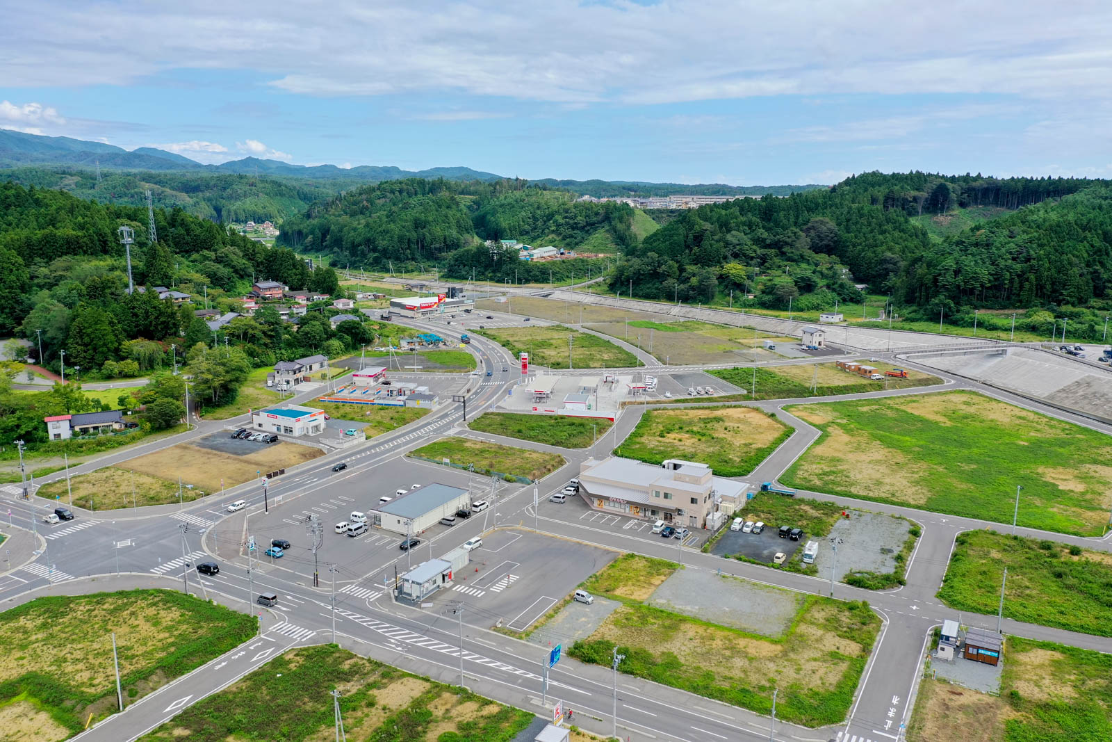 整備された堤防と河川、橋が右手に映り込む市街地の一部を捉え、前景には新しい道路網と小規模な商業施設や駐車場が整備された区画が広がり、背景には田畑と緑豊かな山々が遠くまで連なっている、再開発や復興の進捗がうかがえる風景写真